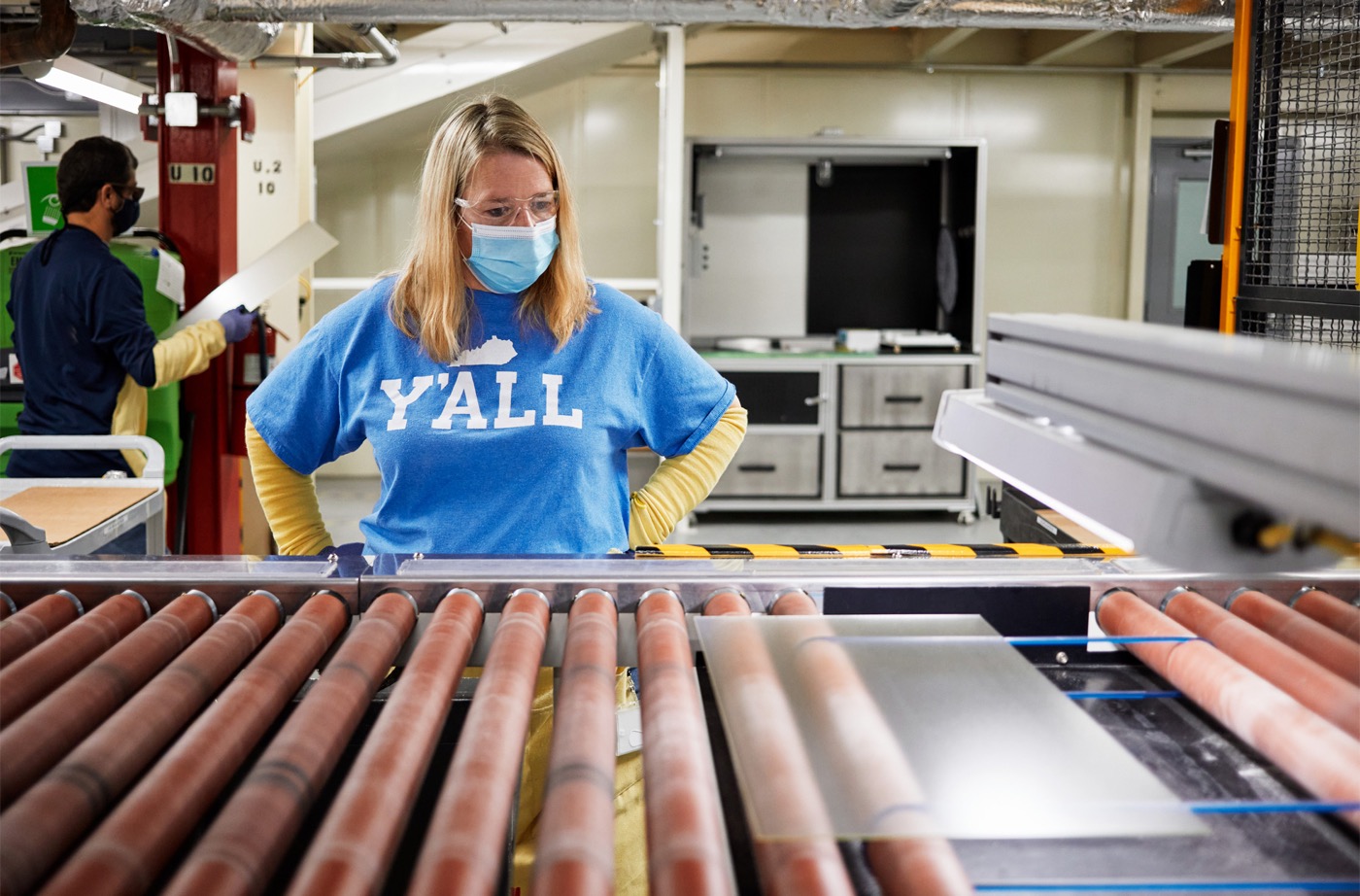  A woman in a blue shirt and face mask looks over a conveyor belt of glass sheets, with another person in the background wearing a white lab coat.
